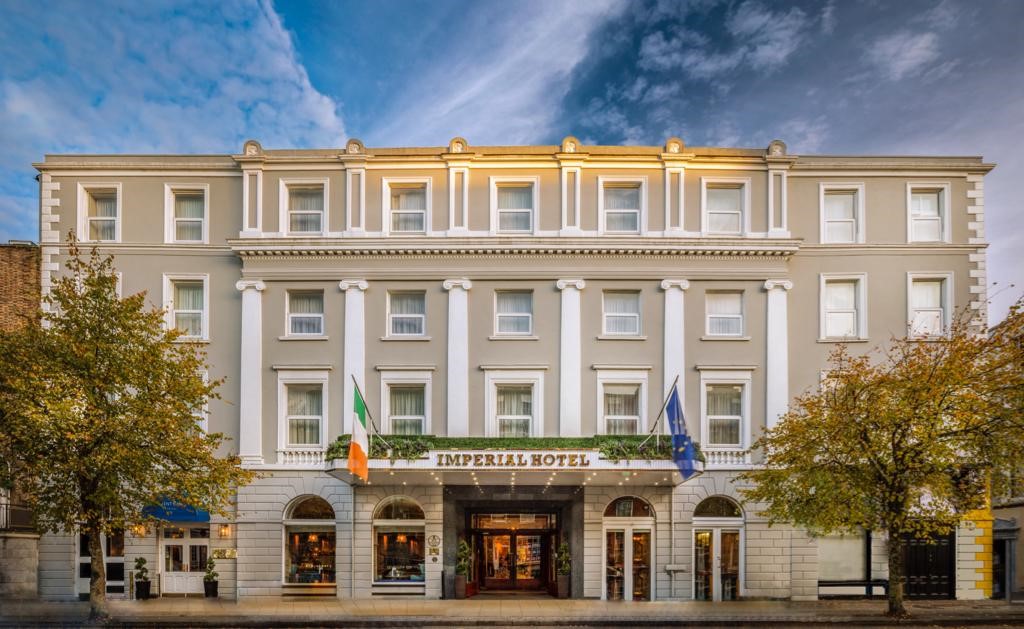 The exterior of The Imperial Hotel on South Mall in Cork, with its cream fa&ccedil;ade, arched entrance, Irish and EU flags, and trees framing the street.